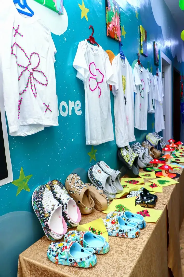 Handmade crafts on a table beneath decorated shirts displayed on a wall